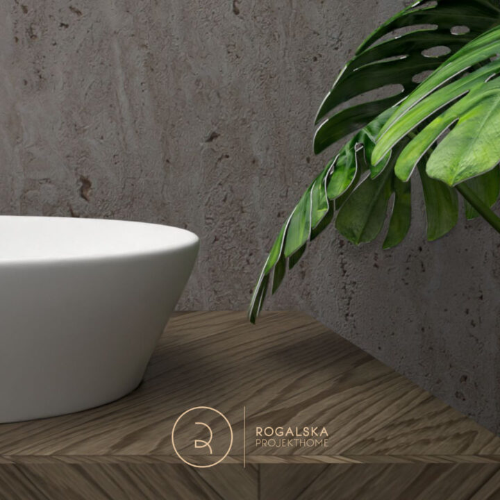 Biała umywalka nablatowa dopełniona kolorami ziemi w postaci blatu z brązowego drewna, szarego kamienia na ścianie oraz roślinności.