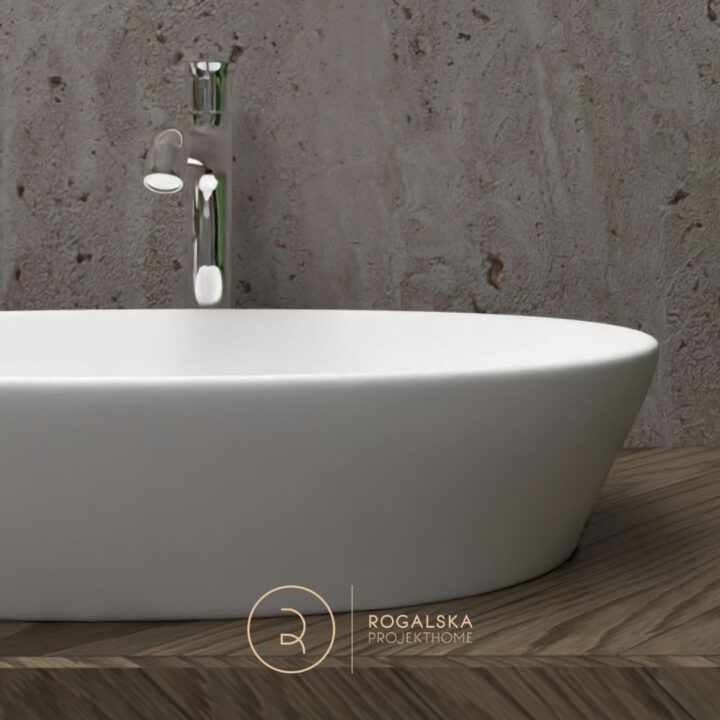 Umywalka nablatowa w kolorze białym, umieszczona na blacie z drewna w jodełkę. Na ścianie szary kamień, który nadaje surowości całemu wnętrzu.