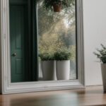 Lustro w szarej ozdobnej ramie, które odbija zielone drzwi oraz widok na świat pełen zielonych, gęstych drzew. Na podłodze stoją donice w kolorze białym z bujną roślinnością.